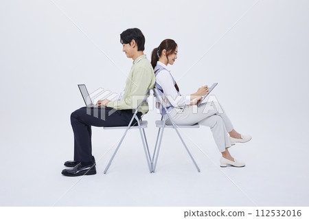 A man and a woman sitting back to back and playing a tablet with a laptop 112532016