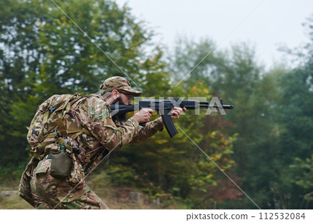An elite soldier, camouflaged and stealthily navigating through dangerous woodland terrain, executes a covert mission in a secluded forest area 112532084