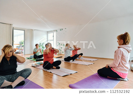 A group of senior women engage in various yoga exercises, including neck, back, and leg stretches, under the guidance of a trainer in a sunlit space, promoting well-being and harmony 112532192