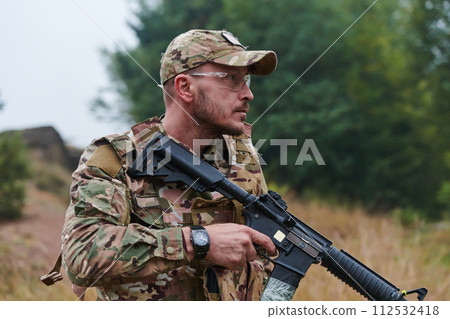 An elite soldier, camouflaged and stealthily navigating through dangerous woodland terrain, executes a covert mission in a secluded forest area 112532418