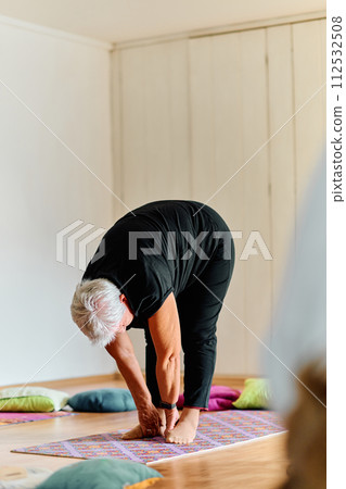 In a sunlit space, a senior woman gracefully practices rejuvenating yoga, focusing on neck, back, and leg stretches, embodying serenity and well-being 112532508