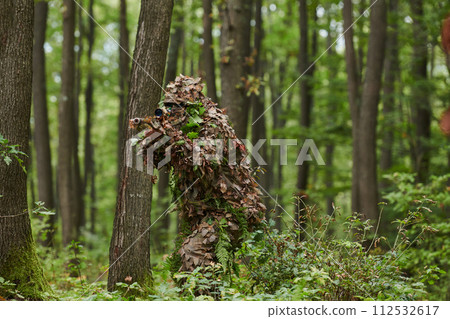 A highly skilled elite sniper, camouflaged in the dense forest, stealthily maneuvers through dangerous woodland terrain on a covert and precise mission 112532617