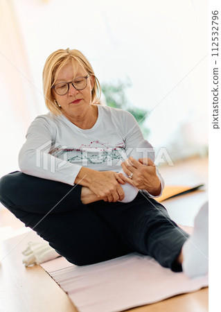 In a sunlit space, a senior woman gracefully practices rejuvenating yoga, focusing on neck, back, and leg stretches, embodying serenity and well-being 112532796