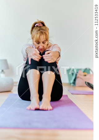 In a sunlit space, a senior woman gracefully practices rejuvenating yoga, focusing on neck, back, and leg stretches, embodying serenity and well-being 112533419