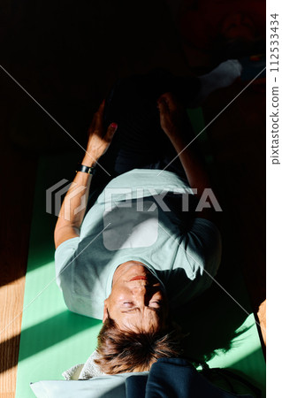 In a sunlit space, a senior woman gracefully practices rejuvenating yoga, focusing on neck, back, and leg stretches, embodying serenity and well-being 112533434