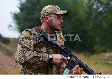 An elite soldier, camouflaged and stealthily navigating through dangerous woodland terrain, executes a covert mission in a secluded forest area 112533784