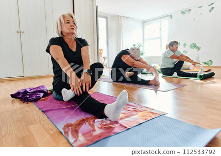 A group of senior women engage in various yoga exercises, including neck, back, and leg stretches, under the guidance of a trainer in a sunlit space, promoting well-being and harmony 112533792
