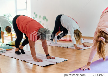 A group of senior women engage in various yoga exercises, including neck, back, and leg stretches, under the guidance of a trainer in a sunlit space, promoting well-being and harmony 112533929