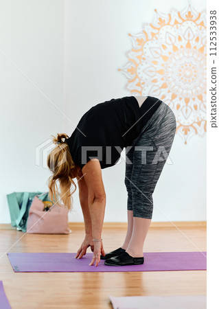 In a sunlit space, a senior woman gracefully practices rejuvenating yoga, focusing on neck, back, and leg stretches, embodying serenity and well-being 112533938
