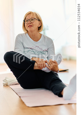 In a sunlit space, a senior woman gracefully practices rejuvenating yoga, focusing on neck, back, and leg stretches, embodying serenity and well-being 112534034
