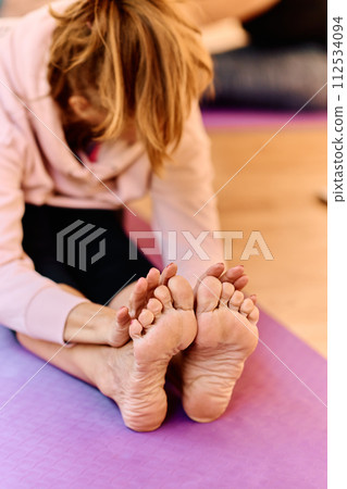 In a sunlit space, a senior woman gracefully practices rejuvenating yoga, focusing on neck, back, and leg stretches, embodying serenity and well-being 112534094
