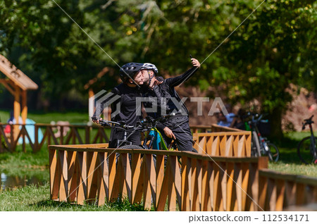 A blissful couple, adorned in professional cycling gear, enjoys a romantic bicycle ride through a park, surrounded by modern natural attractions, radiating love and happiness 112534171