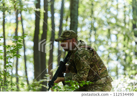 An elite soldier, camouflaged and stealthily navigating through dangerous woodland terrain, executes a covert mission in a secluded forest area 112534490
