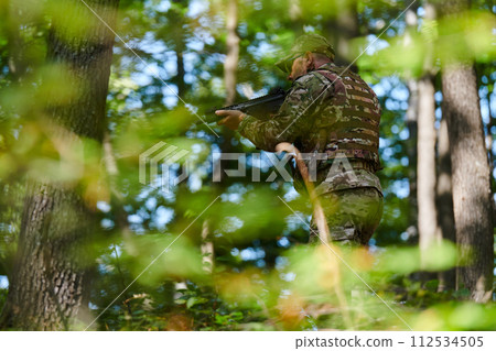 An elite soldier, camouflaged and stealthily navigating through dangerous woodland terrain, executes a covert mission in a secluded forest area 112534505