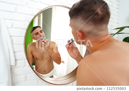 Handsome young man shaving face with electric razor on white background 112535003