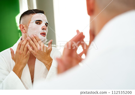 Handsome young man applying sheet mask near mirror in bathroom Handsome young man applying sheet mask near mirror in bathroom 112535200