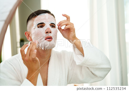 Handsome young man applying sheet mask near mirror in bathroom 112535234