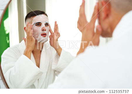 Handsome young man applying sheet mask near mirror in bathroom 112535737