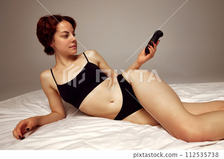 Young woman in black underwear lying on bed, holing vibrator against beige studio background 112535738