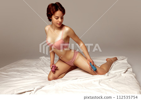 Young woman in underwear relaxing on bed, holding vibrator toy against beige studio background Young woman in underwear relaxing on bed, holding vibrator toy against beige studio background 112535754