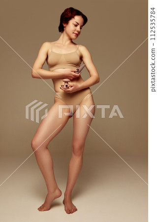 Full-length image of young woman with slim body, standing in underwear, holding various vibrators against beige studio background Full-length image of young woman with slim body, standing in underwear, holding various vibrators against beige studio background 112535784