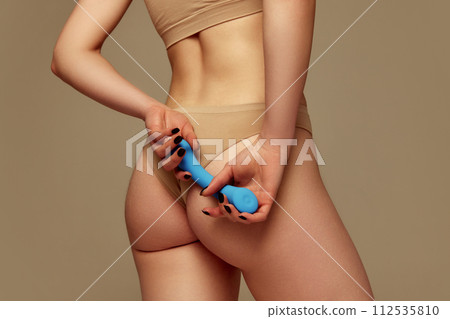 Cropped image of female body, buttocks in underwear. Woman holding blue vibrator toy against beige studio background Cropped image of female body, buttocks in underwear. Woman holding blue vibrator toy against beige studio background 112535810