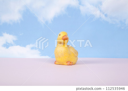 a plaster figure of a yellow chick on a pink ground and in front of a blue sky background with clouds a plaster figure of a yellow chick on a pink ground and in front of a blue sky background with clouds 112535946