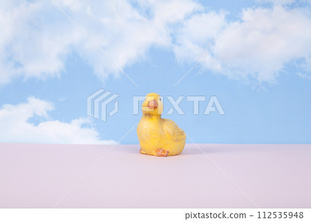 a plaster figure of a yellow chick on a pink ground and in front of a blue sky background with clouds 112535948