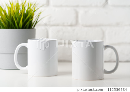 Two mugs on white table against brick wall background 112536584
