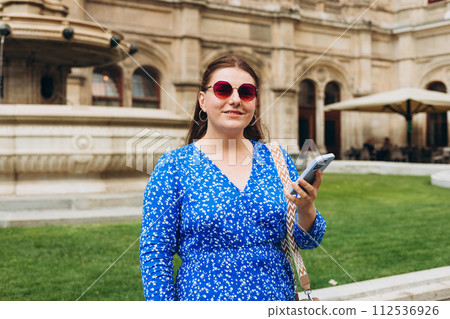 Happy cheerful young woman in sunglasses walking on city street checks her smartphone. Portrait of beautiful 30s girl using smartphone outdoors. Urban lifestyle concept, summer time 112536926