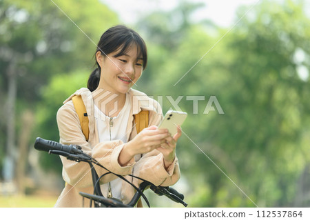 Attractive young woman standing near a bicycle at a city park and using a mobile phone 112537864
