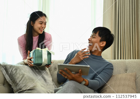 Beautiful young woman making a surprise giving a present to beloved husband at home Beautiful young woman making a surprise giving a present to beloved husband at home 112537945