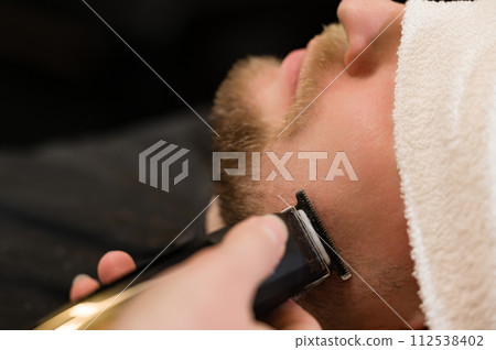 Cutting a gentlemans beard in a barbershop with a clipper. Shortening the length of the beard from the sides by the master for the client. Cutting a gentlemans beard in a barbershop with a clipper. Shortening the length of the beard from the sides by the master for the client. 112538402