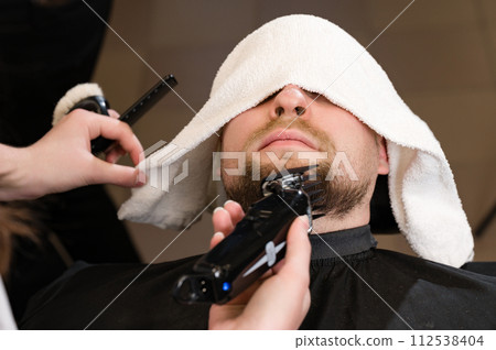 The clients head is covered with a towel during a beard trim. Shortening the length of the beard from the sides by the master for the client. 112538404