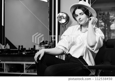 Portrait of a young hairdresser girl in a hat. A female barber is sitting in a chair and waiting for a client. Portrait of a young hairdresser girl in a hat. A female barber is sitting in a chair and waiting for a client. 112538450
