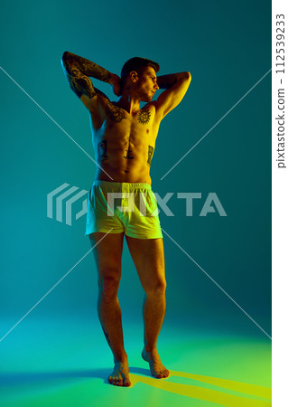 Young man with muscular, athletic body posing raising arms holding behind head in neon light against gradient studio background. 112539233