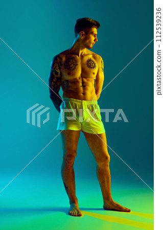 Portrait of tattooed young handsome man with muscular body looking away in neon light against gradient studio background. 112539236