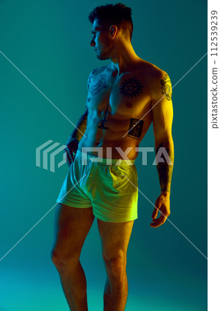 Young athletic man with naked torso posing in neon light against gradient studio background. Male model with tattoos on body. 112539239