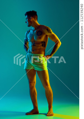 Tattooed, young man posing in underwear holds hands on hips in neon light against gradient studio background. Male model with athletic body. Tattooed, young man posing in underwear holds hands on hips in neon light against gradient studio background. Male model with athletic body. 112539240