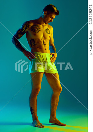 Young attractive shirtless man with tattoos on his athletic body posing in underwear in neon light against gradient studio background. 112539241