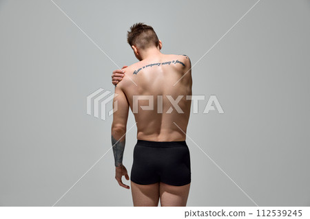 Rear view of young man with tattoos on body holds hand on shoulder shows pain against grey studio background. Copy space. 112539245
