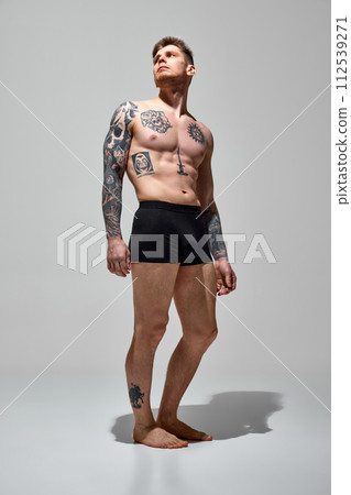 Young attractive shirtless man with tattoos on his athletic body posing in underwear against grey studio background. Young attractive shirtless man with tattoos on his athletic body posing in underwear against grey studio background. 112539271