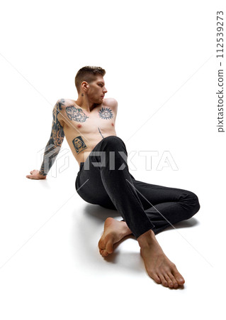 Tattooed, handsome man wearing black pants sitting, posing against white studio background. Copy space. Tattooed, handsome man wearing black pants sitting, posing against white studio background. Copy space. 112539273