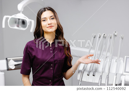 A young pretty female dentist is standing near the dental chair in the office, gesturing with her hands A young pretty female dentist is standing near the dental chair in the office, gesturing with her hands 112540092