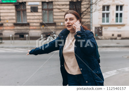 Stylish 30s woman with backpack catching a taxi. Woman doing multiple tasks. Middle age woman talking on smartphone and calling taxi cab 112540157