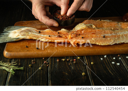 The cook uses his hands to add dry aromatic spices to raw fish fillets on the kitchen table. Concept of preparing fish herring in restaurant kitchen 112540454