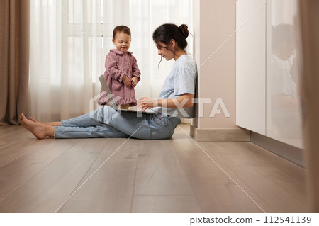 beautiful casual woman sitting on the floor and working on laptop with her little child girl at home beautiful casual woman sitting on the floor and working on laptop with her little child girl at home 112541139