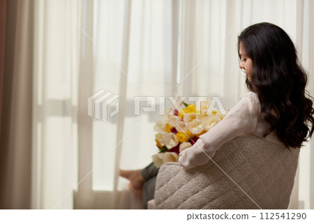 Beautiful woman with spring flowers tulips in hands sitting on chair near window. Women's Day Beautiful woman with spring flowers tulips in hands sitting on chair near window. Women's Day 112541290