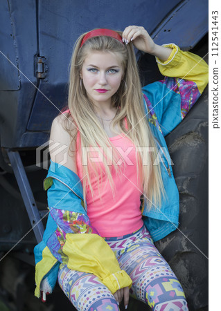 A beautiful country girl in the style of the 90s in bright clothes stands near a tractor. 112541443