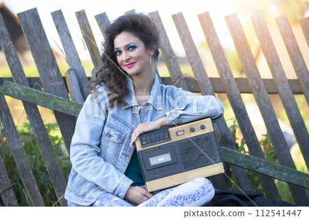 A beautiful country girl in bright clothes sits on a wooden bench with a cassette recorder. Woman in the style of the 90s. 112541447
